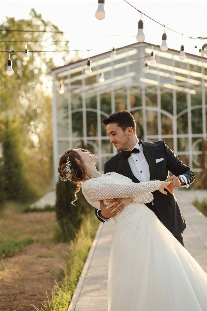A groom dips his bride in front of a glass pavilion hung with string lights