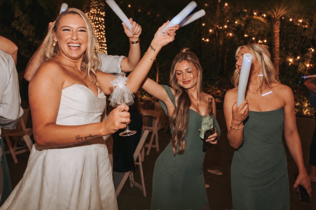 A bride laughing and dancing with two bridesmaids in green dresses under string lights