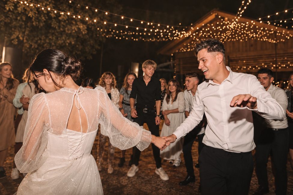 A bride and groom lead a group dance while wedding guests join in under string lights